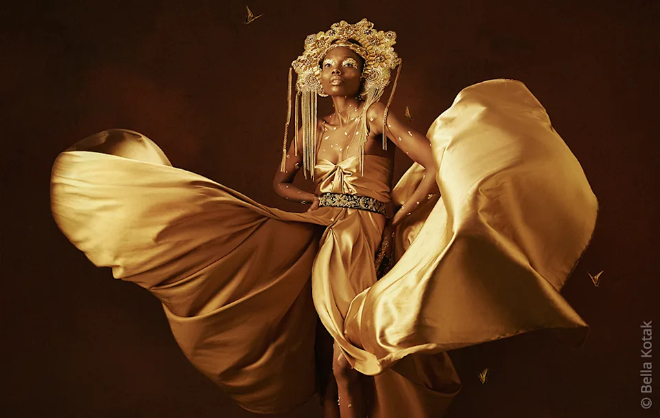 LP | CDP | Magazin | Image Grid Section | Inspiration | Image Person in a gold-colored dress with fabric panels, wearing an ornate headdress, slightly red background.