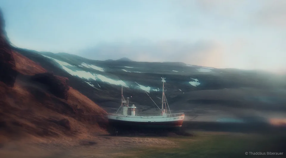 An old fishing boat stands on a wooded shore, surrounded by mountains.